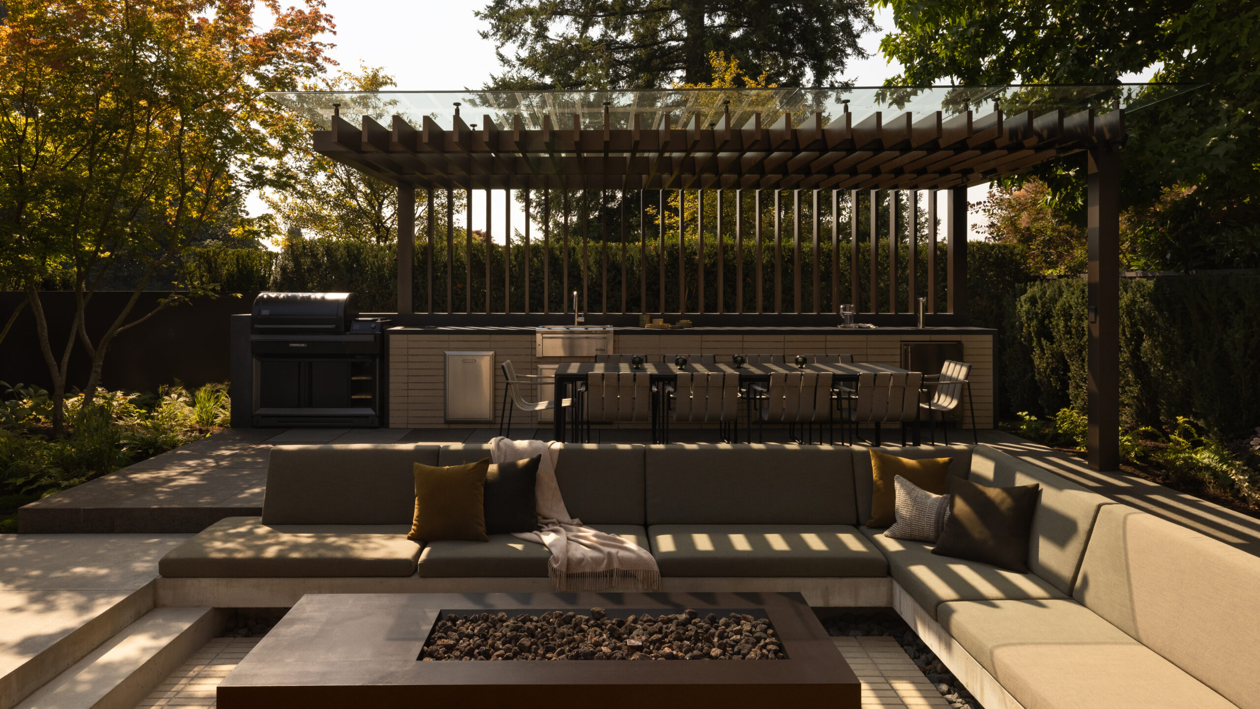 Modern outdoor kitchen and dining area under a slatted pergola with a built-in grill and long dining table, surrounded by trees and shrubs.