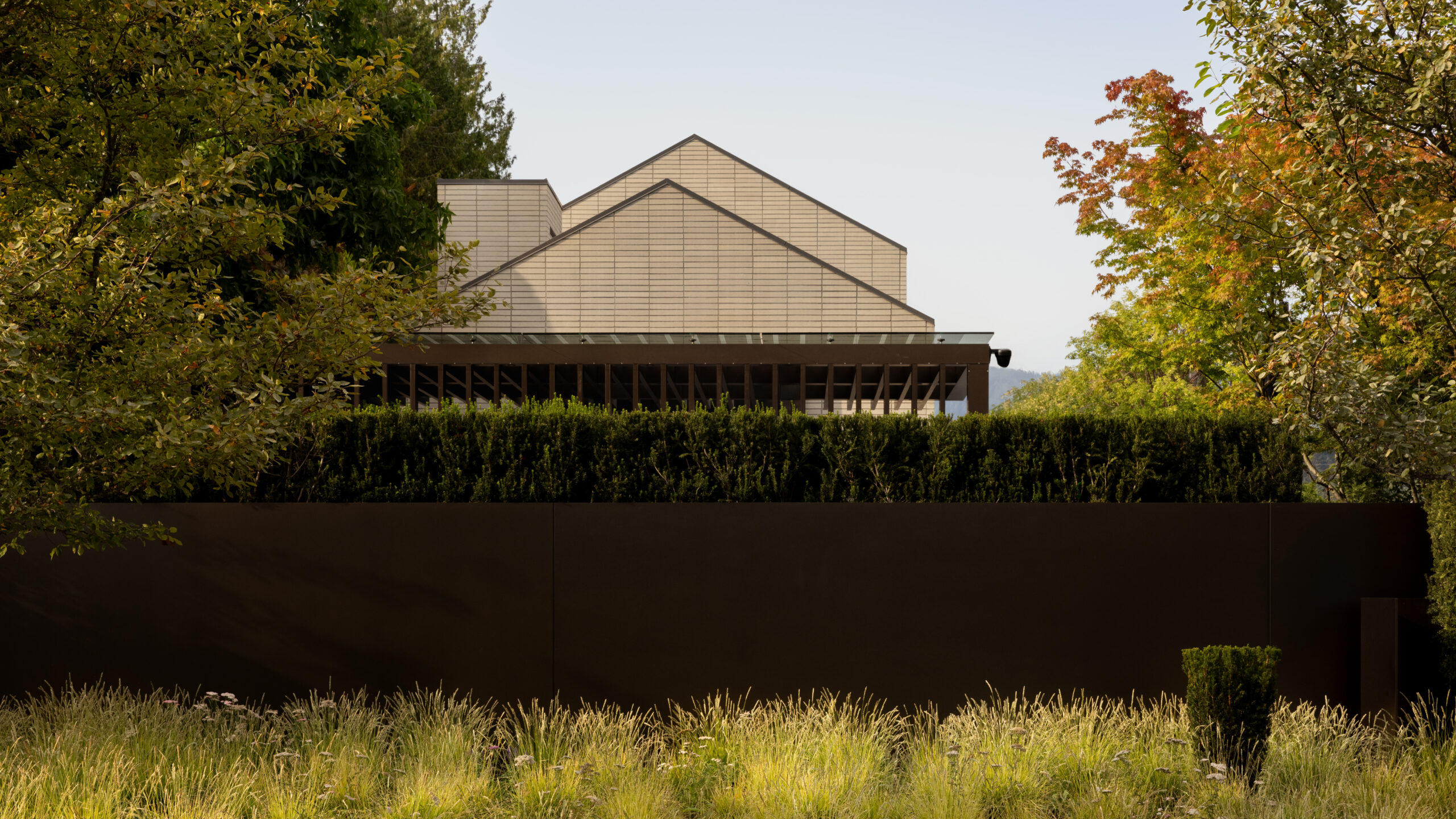 Low-angle view of a modern beige-brick building with triangular roof peaks, framed by green trees and a tall hedge in the foreground.