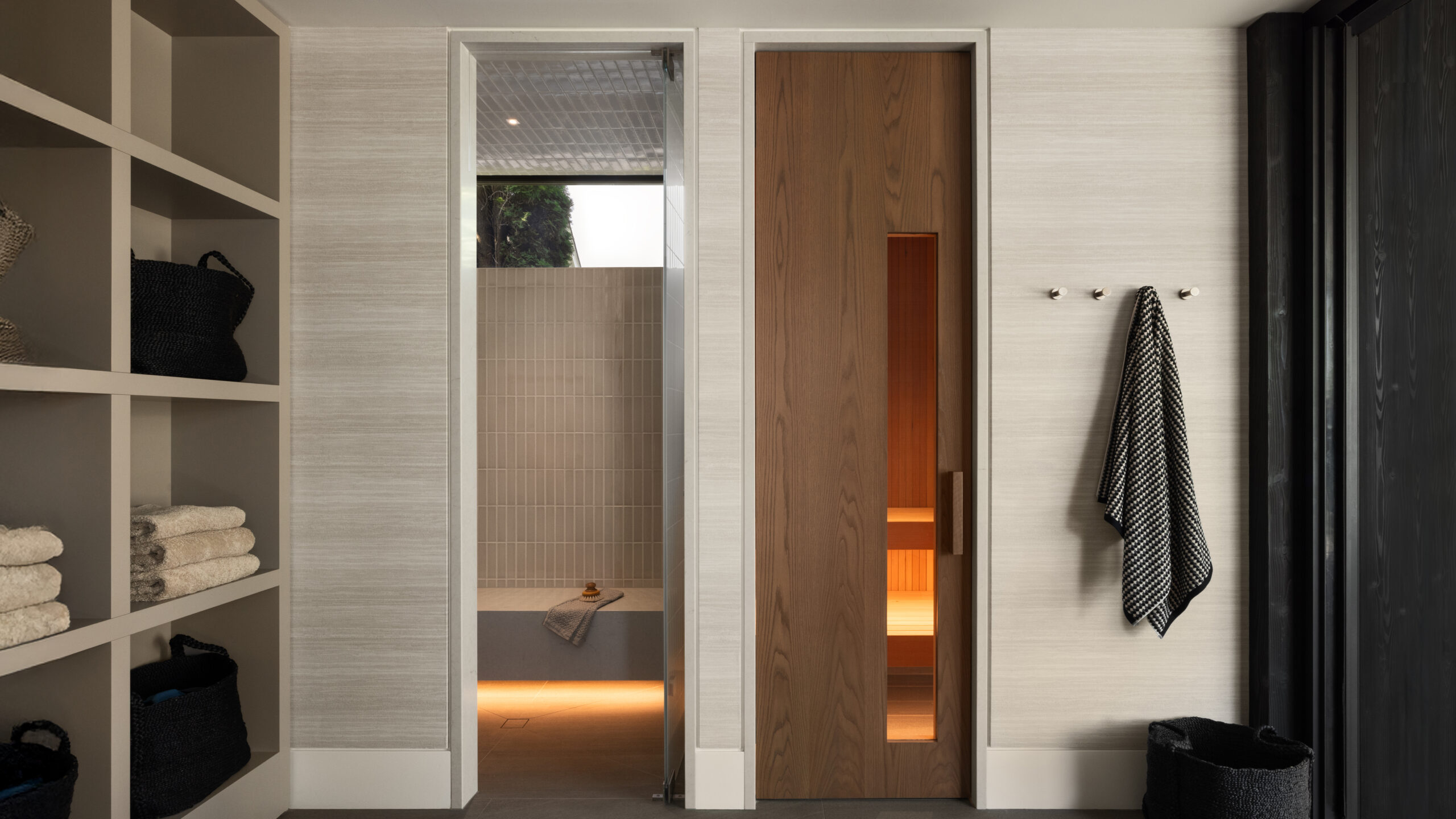 Open shelving with towels and baskets on the left, a tiled shower visible through a central doorway, and a tall wood door on the right.
