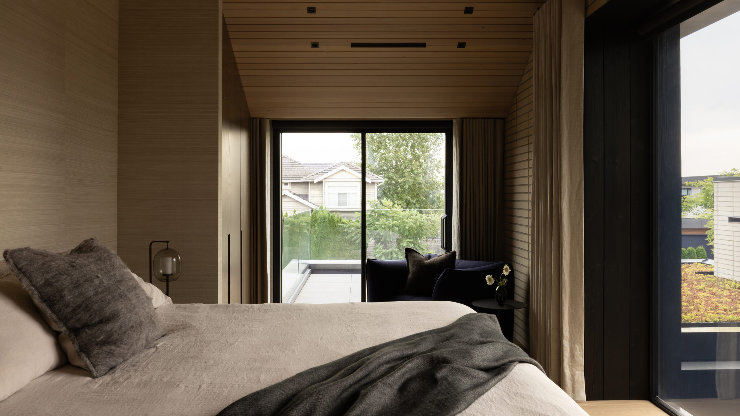 Bedroom with wood panel walls and a large sliding glass door opening to a balcony; outside, trees and a neighboring house are visible.