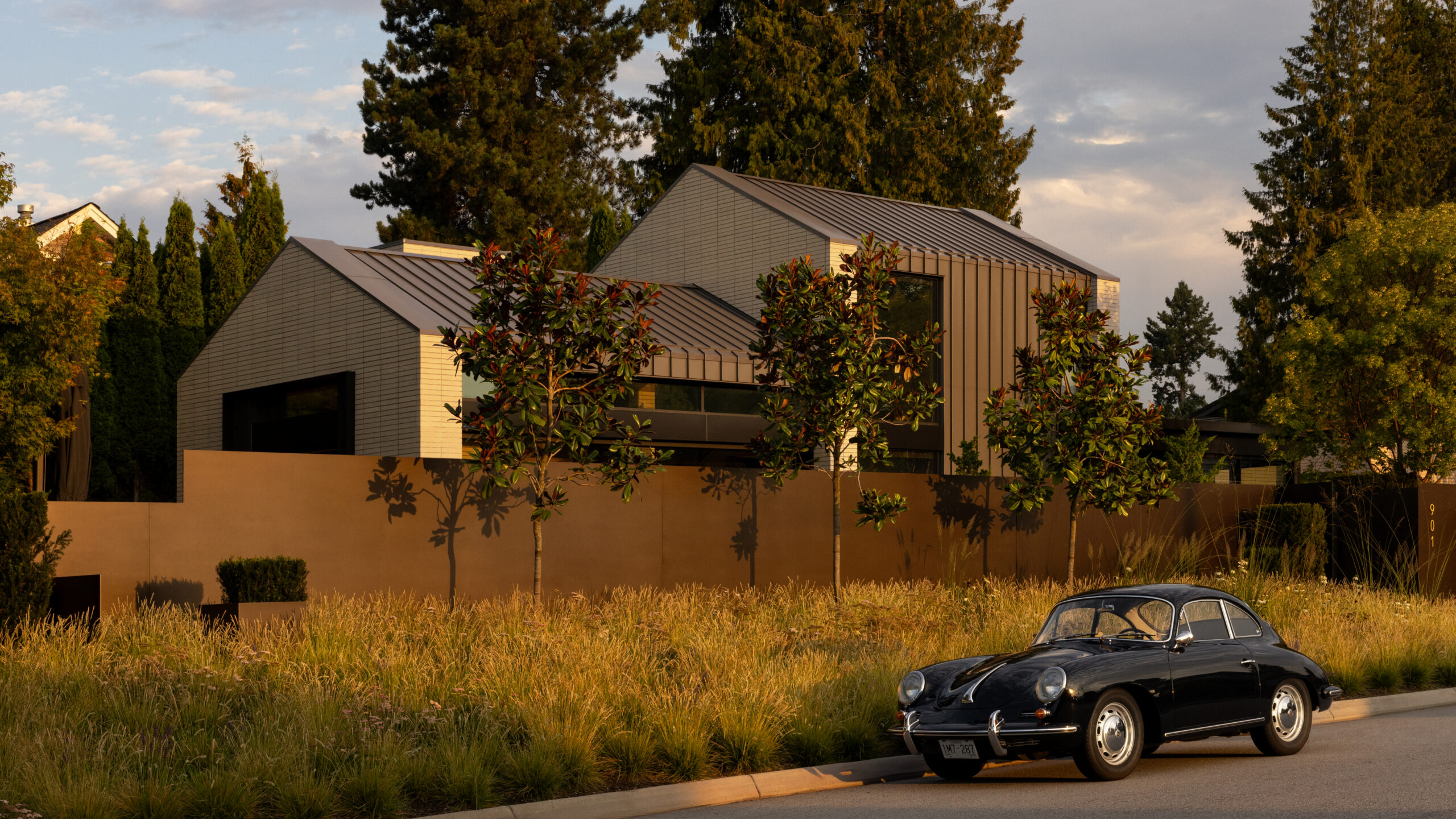 Modern angular house with metal siding and large windows, surrounded by trees at golden hour, with a classic black car parked on the curb.