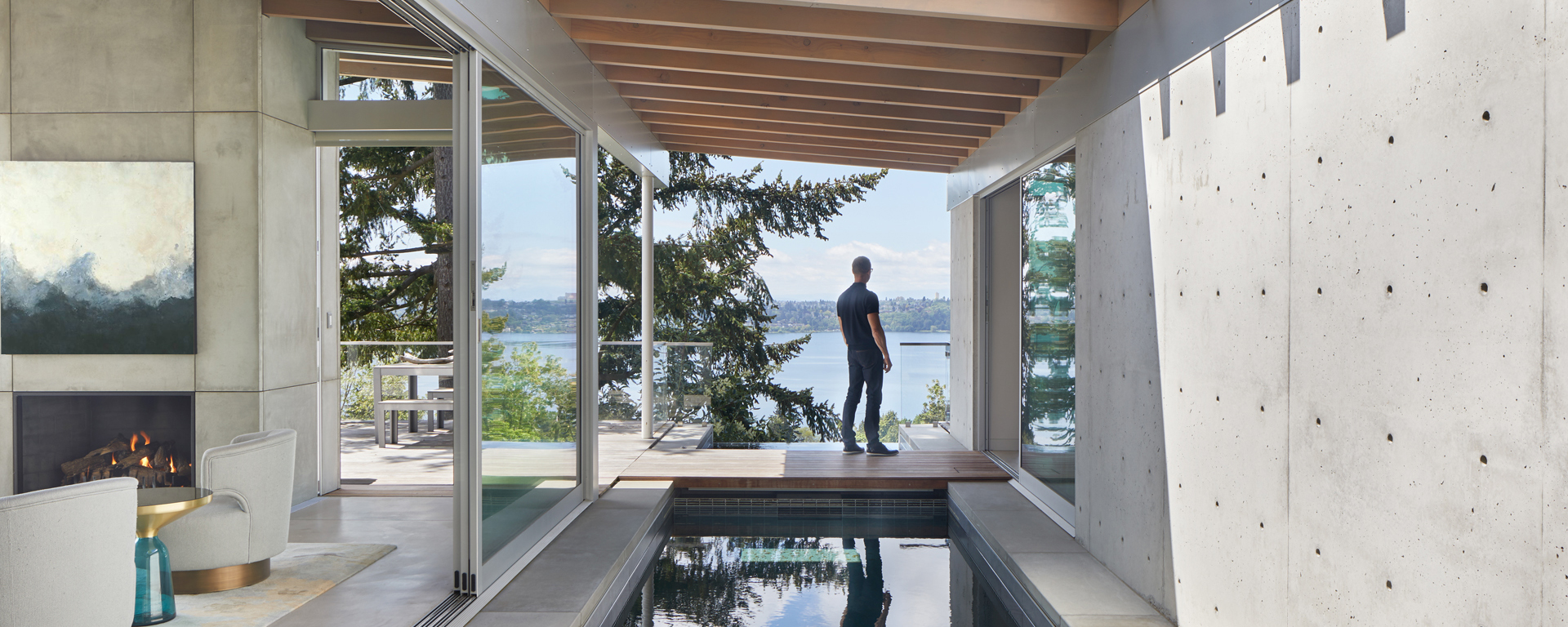 Man stands on a wooden deck gazing at a lake through large glass doors in a modern living space with a fireplace and seating area to the left.