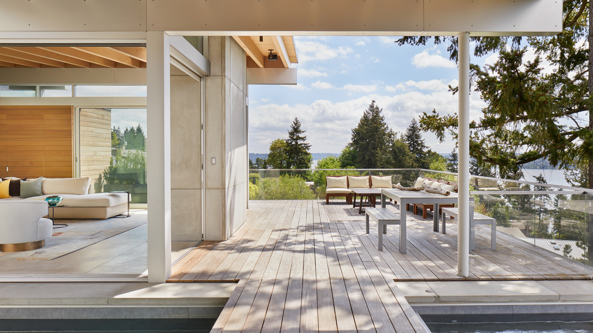 Open-plan living area with beige sectional sofa flowing onto a wooden deck, glass railing and a scenic lake view.